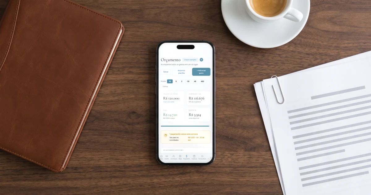 App de orçamento de casamento: celular deitado sobre uma mesa de madeira escura com o orçamento aberto, mostrando o total, o comprometido, o valor pago e o aviso de um pagamento que vence esta semana, ao lado de uma agenda de couro, contratos presos com clipe e um cafezinho