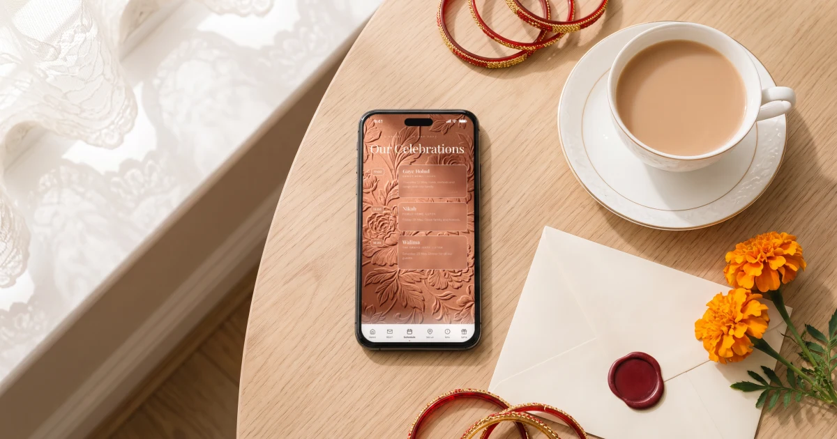 Bilingual Bengali and English wedding invitation for a British-Bangladeshi wedding: a phone lying flat on an oak table in a London home shows the Gaye Holud, Nikah and Walima schedule, beside milky tea, a sealed envelope, glass bangles and marigolds