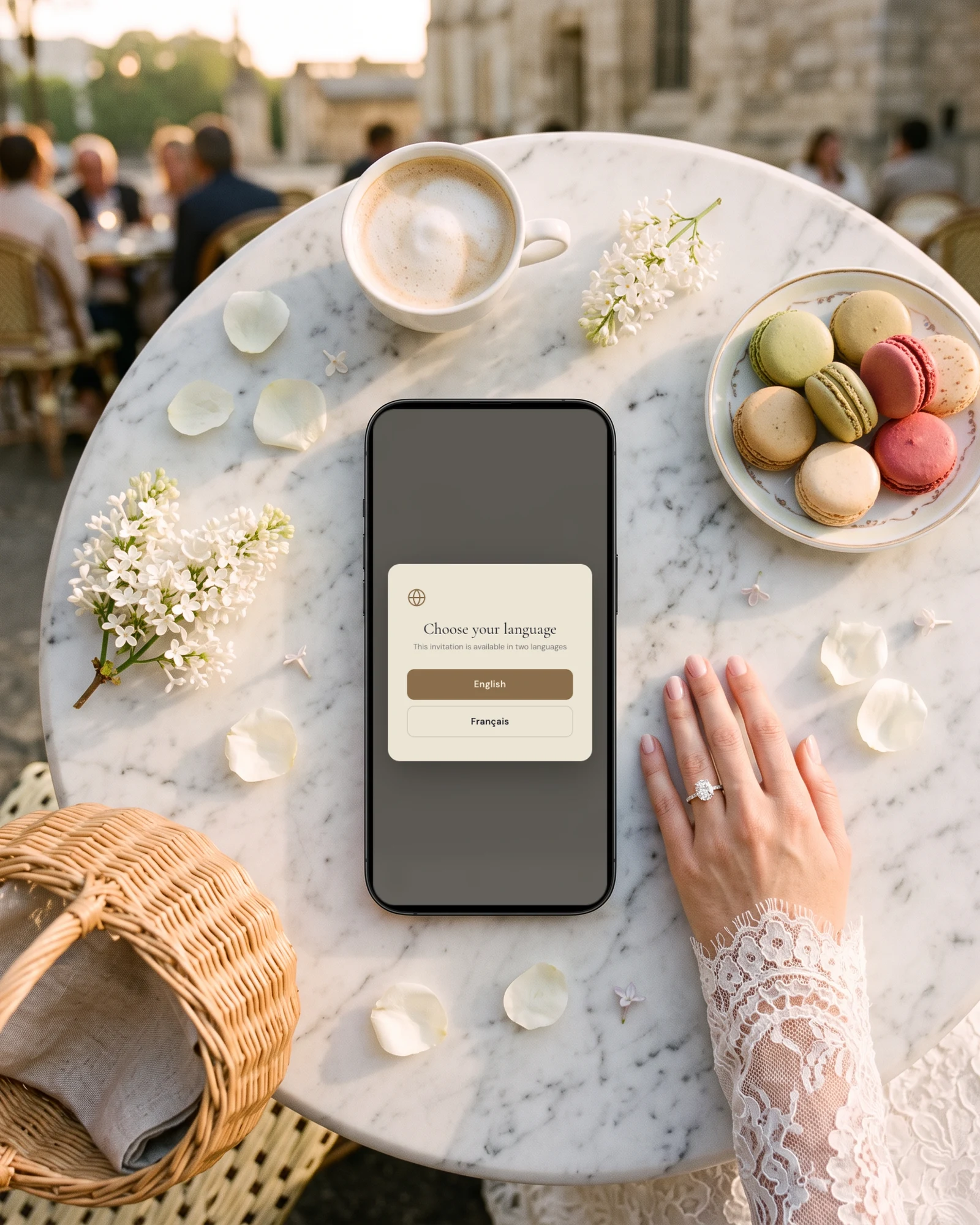 Bilingual wedding invitation in English and French: a phone on a marble Paris cafe table shows the language choice, English or Français, next to macarons, a cafe creme, white lilac and a bride's hand in lace