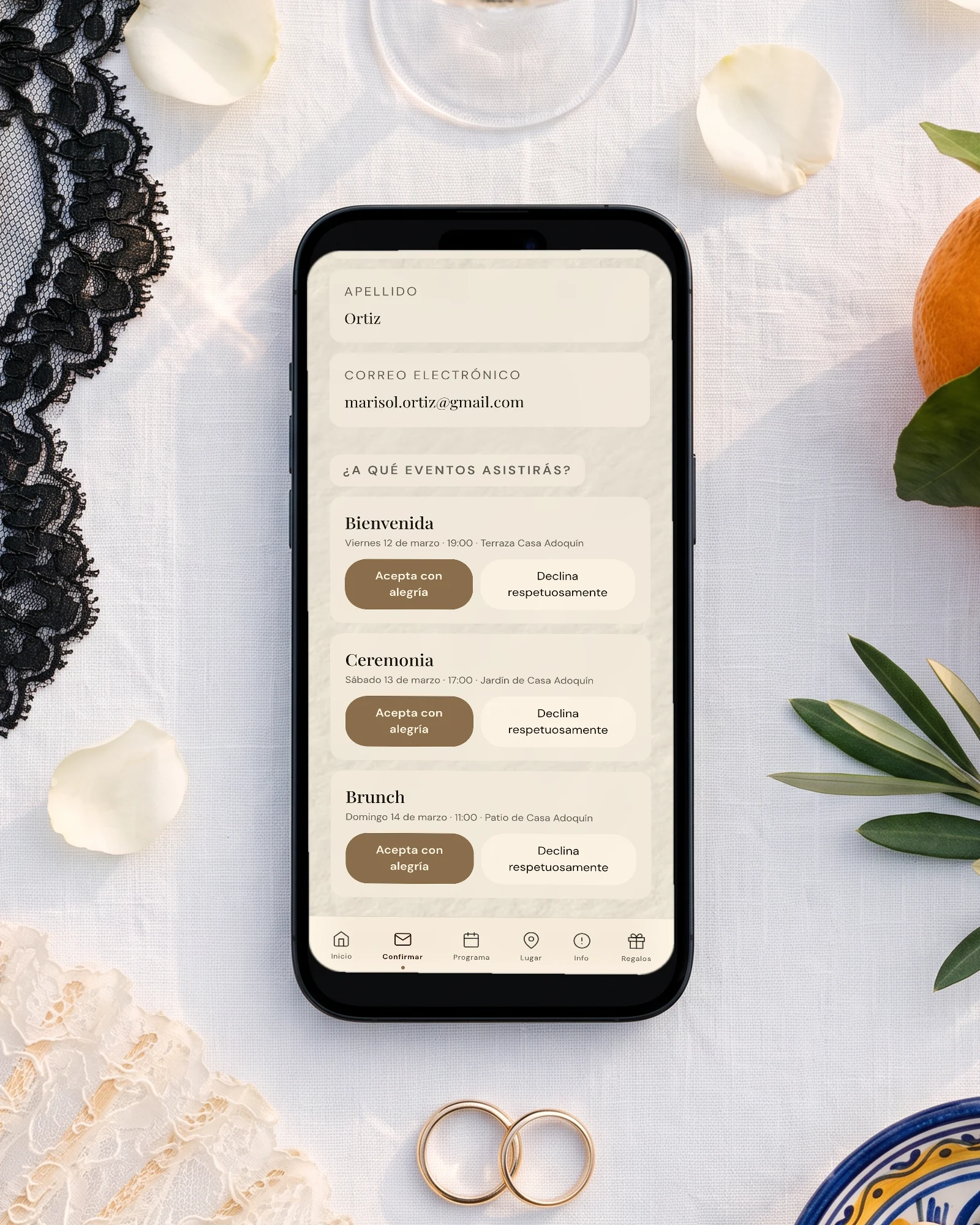 Preboda, boda y postboda en la misma invitación: el teléfono muestra la confirmación por evento con la bienvenida del viernes, la ceremonia del sábado y el brunch del domingo, entre una mantilla de encaje, un abanico, naranjas y una copa de cava