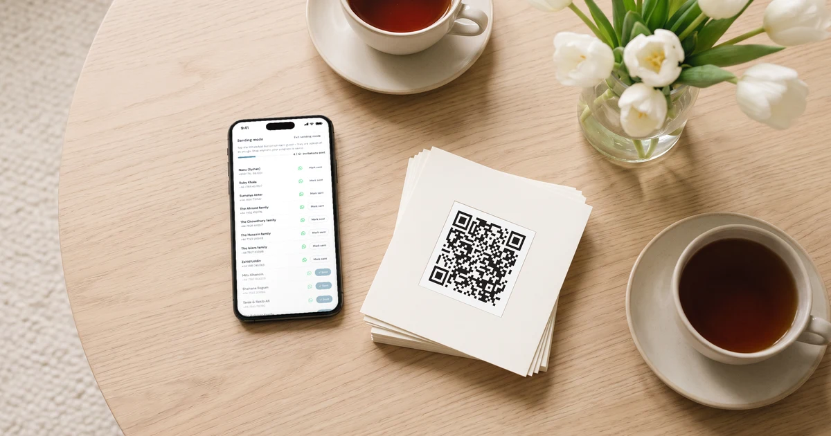 A phone lying on a round wooden table shows sending mode for WhatsApp wedding invitations, a guest list with a sending progress bar, beside a stack of printed QR invitation cards, cups of tea and white tulips