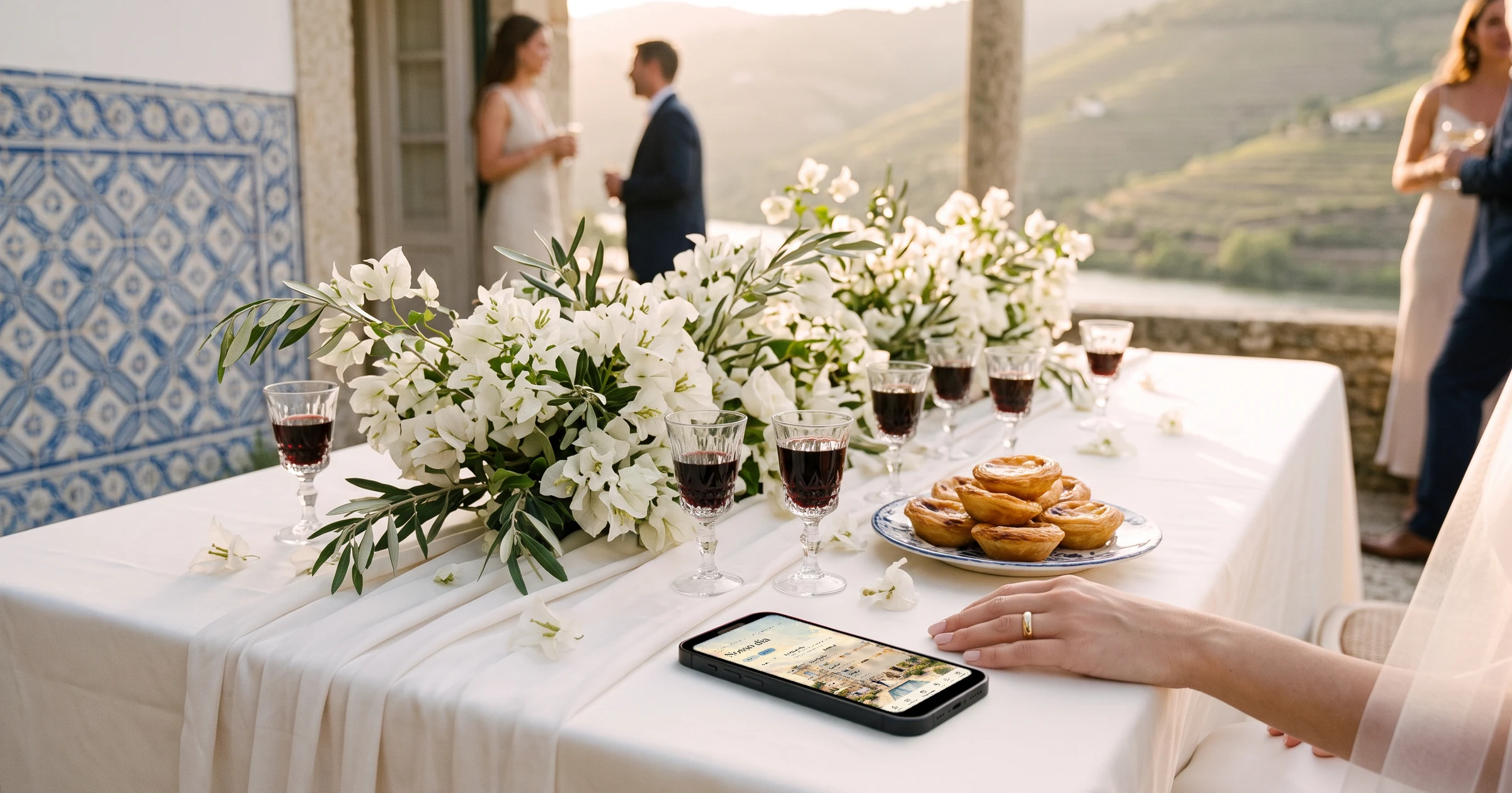 Casamento em Portugal com convidados do Brasil: no terraço de uma quinta no Douro, com azulejos e vinhas ao fundo, um celular sobre a mesa mostra a programação do casamento com abas para o dia 1 e o dia 2, ao lado de pastéis de nata, cálices de vinho do Porto e flores brancas