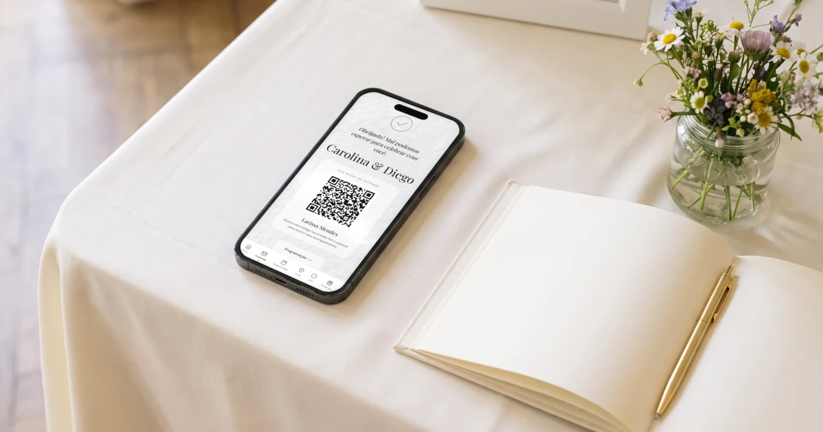 Check-in dos convidados na entrada do casamento: celular deitado sobre a mesa de recepção mostrando o passe de entrada com QR code de uma convidada, logo após a confirmação de presença, ao lado de um livro de assinaturas aberto, uma caneta dourada e um arranjo de flores do campo
