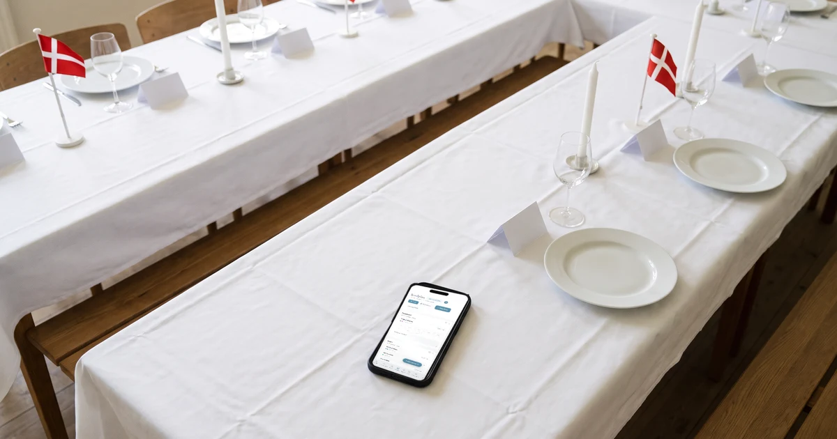 Bordplan bryllup regler: et hesteskobord dækket med hvide duge, lys og små dannebrogsflag, bordkort ved hver kuvert og en telefon, der ligger fladt på bordet og viser bordplanen