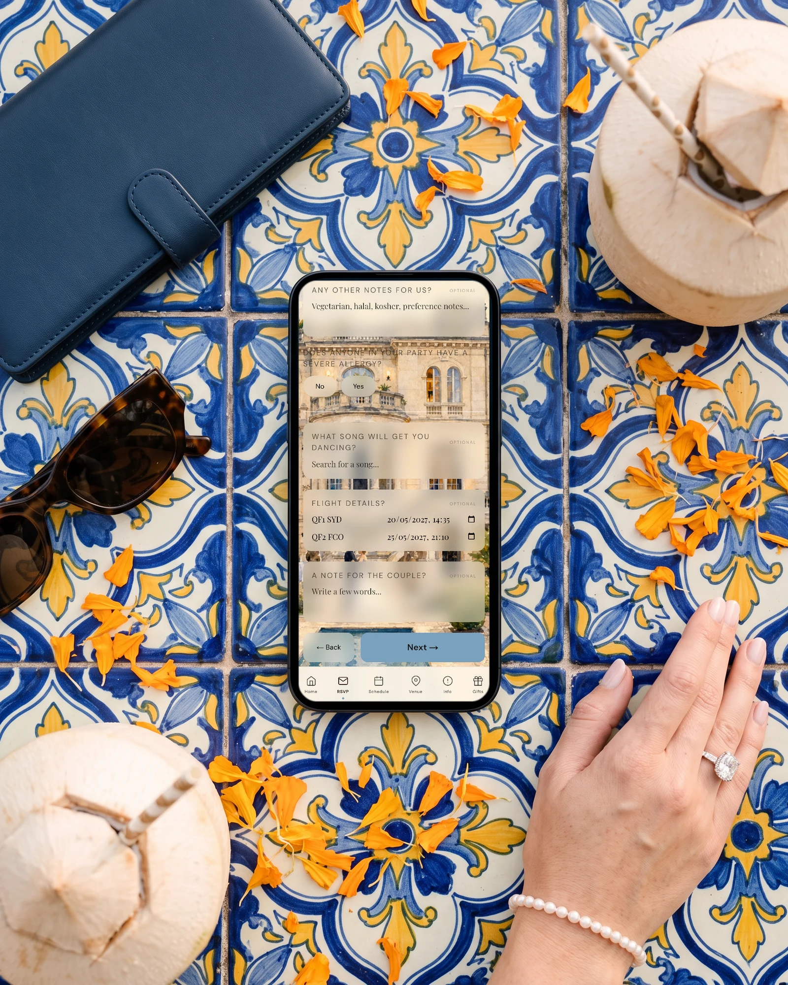 Destination wedding RSVP with guest flight details on a phone lying on talavera tiles: arrival and return flights entered on the form, beside marigold petals, sunglasses, a travel wallet, a coconut and a bride's hand