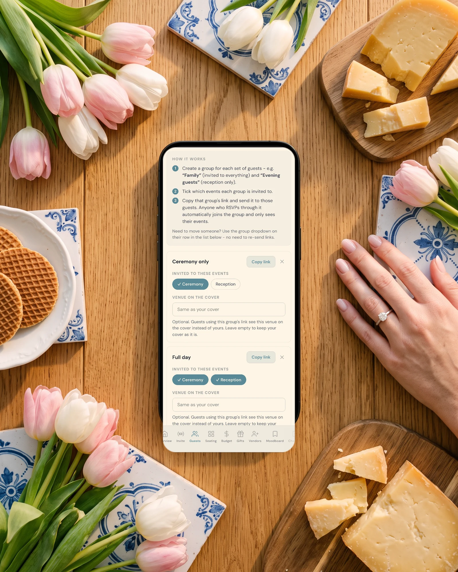 Dutch wedding guest groups on a phone: one group link for ceremony-only guests and one for full-day guests, lying on an oak table with pink and white tulips, Delft blue tiles, stroopwafels, Gouda and a bride's hand