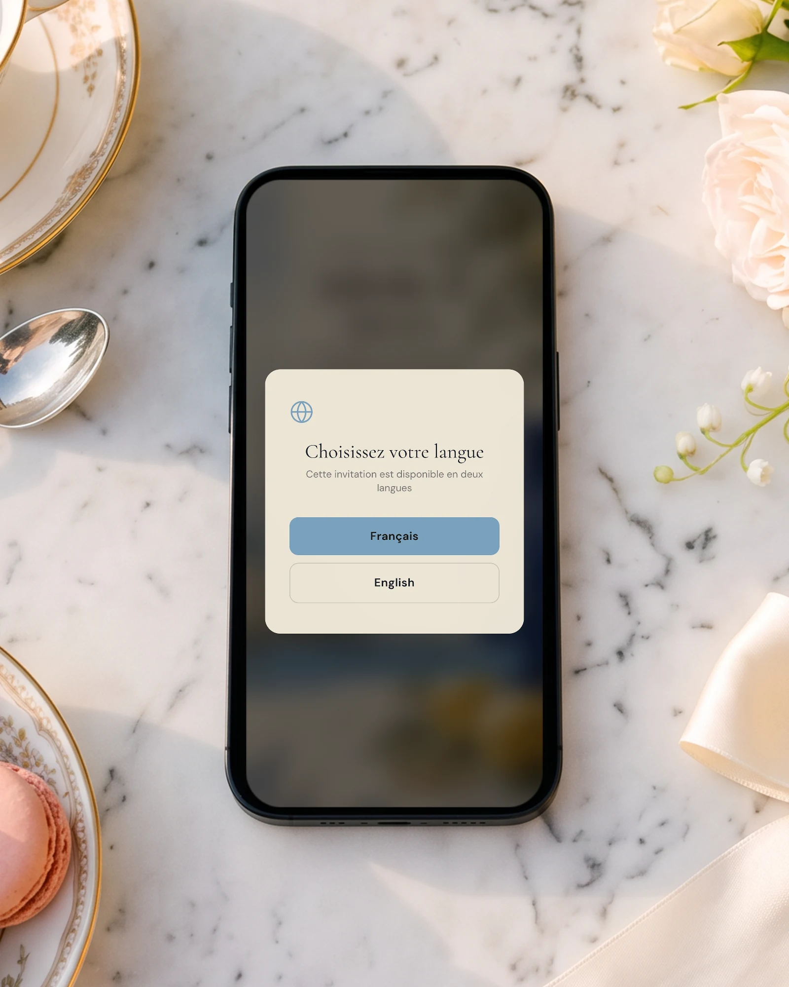 Faire-part bilingue français anglais : un téléphone posé sur une table de marbre, entre une tasse de porcelaine anglaise, des macarons et des roses, affiche l'écran Choisissez votre langue avec Français et English