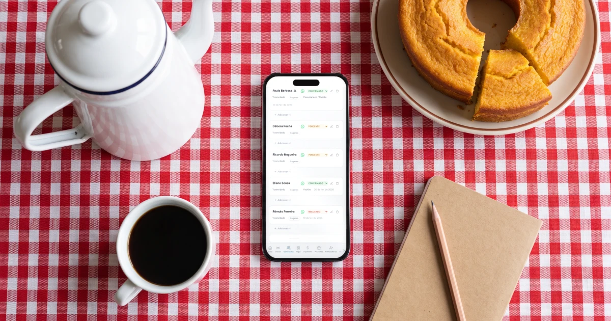Família ajudando na lista de convidados do casamento: celular deitado sobre a toalha xadrez da mesa da cozinha mostrando a lista de convidados com confirmados e pendentes, ao lado de um bule esmaltado, uma xícara de café, um bolo de fubá e um caderno