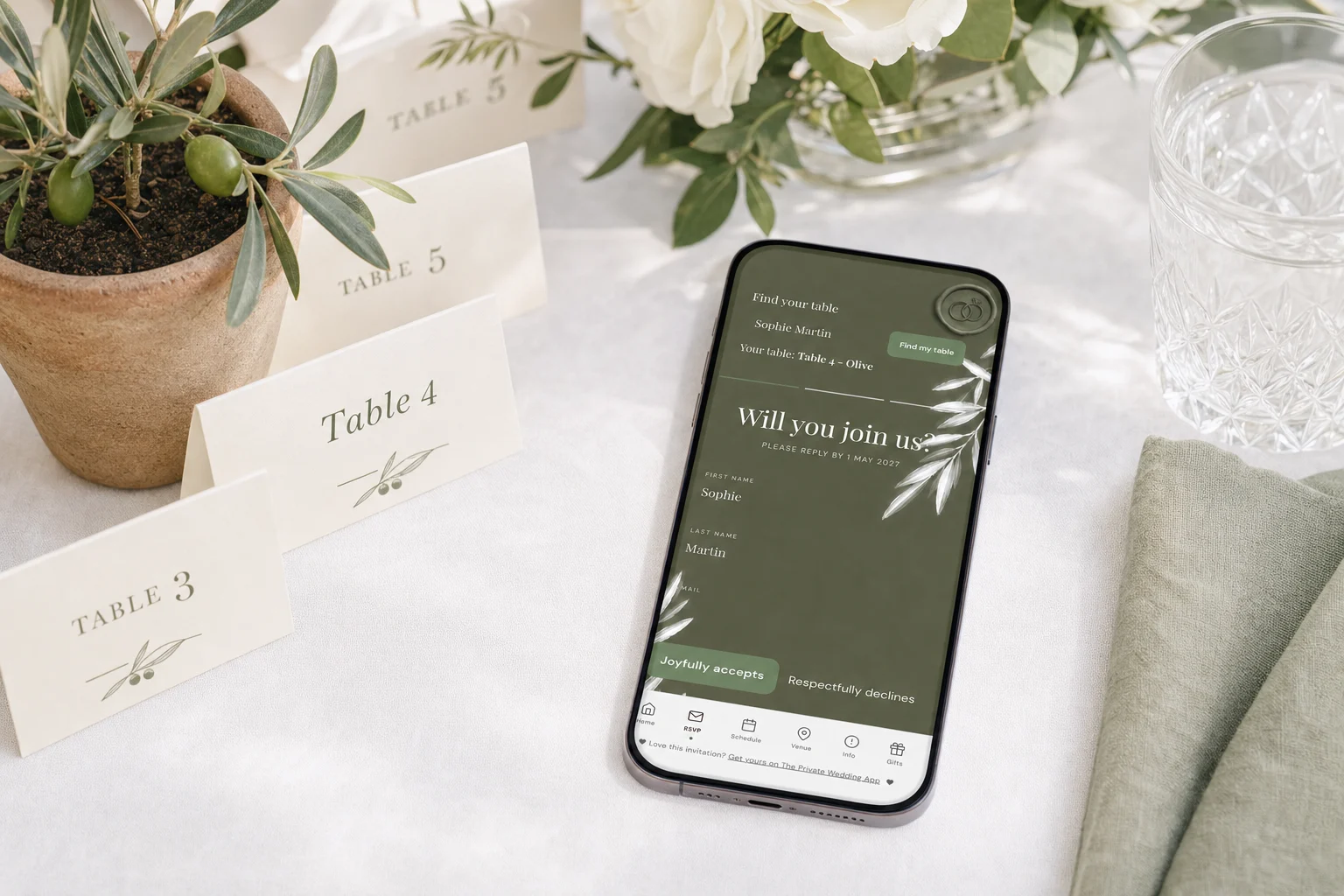 Find your table at a wedding: a phone shows a guest typing her name on the invitation and seeing Table 4, next to escort cards with table numbers on a white reception table