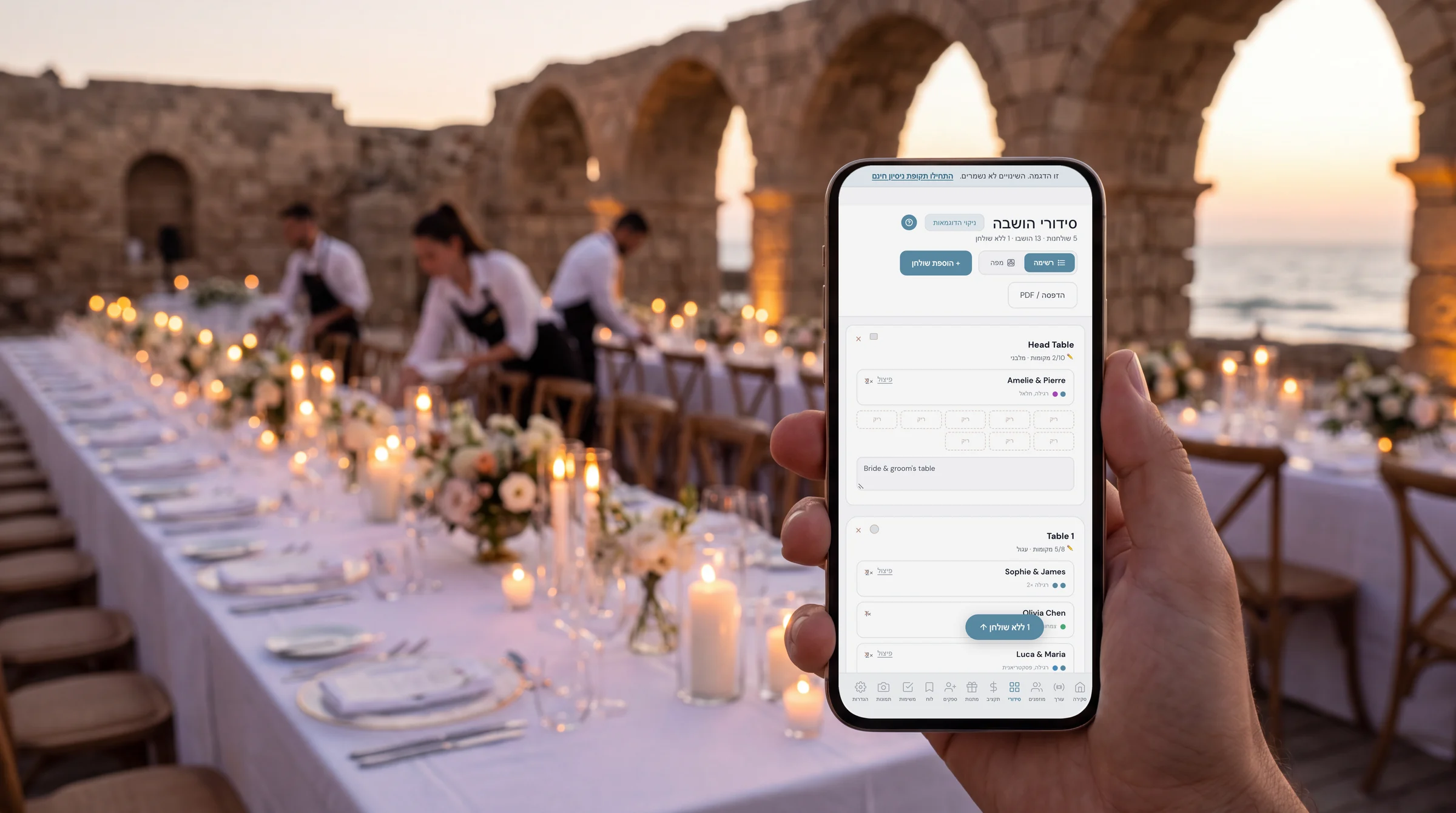 סידורי הושבה לחתונה: יד מחזיקה טלפון עם סידורי ההושבה בעברית, מול שולחן ארוך ערוך עם מפה לבנה ונרות, קשתות אבן עתיקות והים בקיסריה בשעת בין ערביים