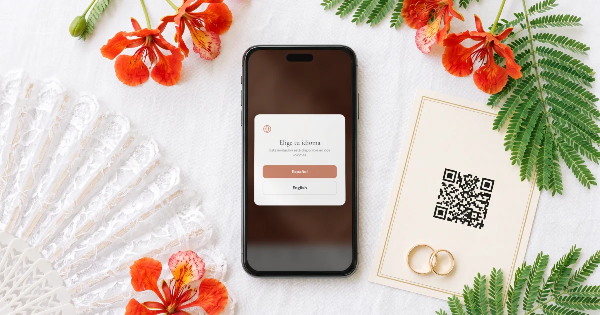 Invitaciones de boda bilingües español-inglés: un teléfono sobre una mesa clara muestra el selector de idioma de la invitación con español e inglés, junto a un abanico de encaje, flores de flamboyán y una tarjeta QR impresa