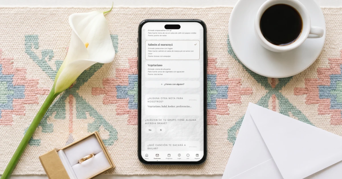 Invitaciones de matrimonio digitales en Colombia: un teléfono acostado muestra la confirmación de asistencia con la elección del menú, junto a una taza de café colombiano, flores de cartucho y un sobre de lluvia de sobres