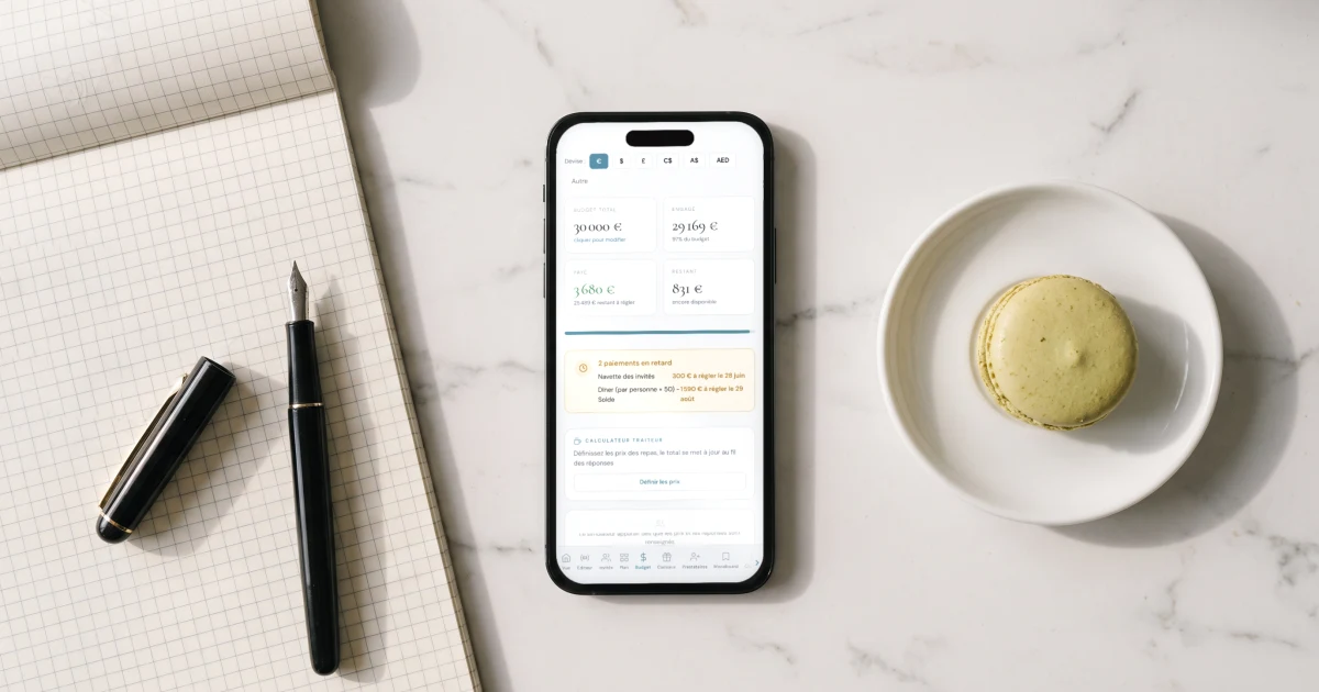 Meilleure application budget mariage : un téléphone affiche le budget du mariage en euros, payé et restant, à côté d'un carnet quadrillé, d'un stylo plume et d'un macaron