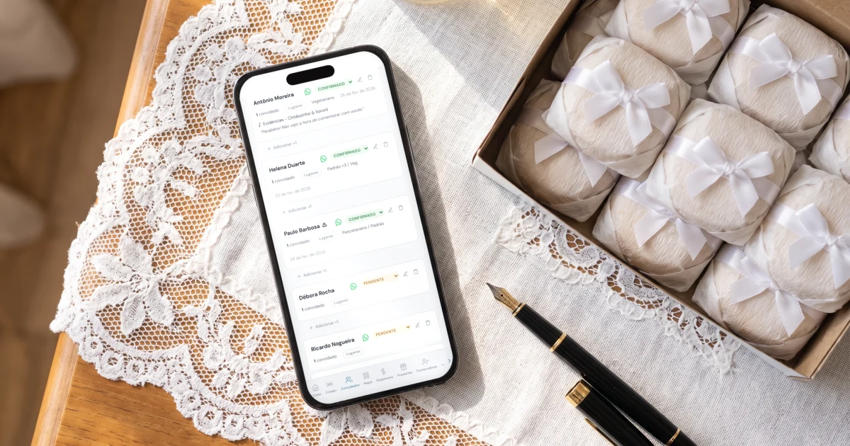 Melhor app de lista de convidados de casamento: celular deitado sobre uma toalha de renda com a lista de convidados aberta, mostrando confirmados, pendentes e recusados, o prato de cada convidado e o botão de WhatsApp, ao lado de uma caixa de bem-casados e uma caneta
