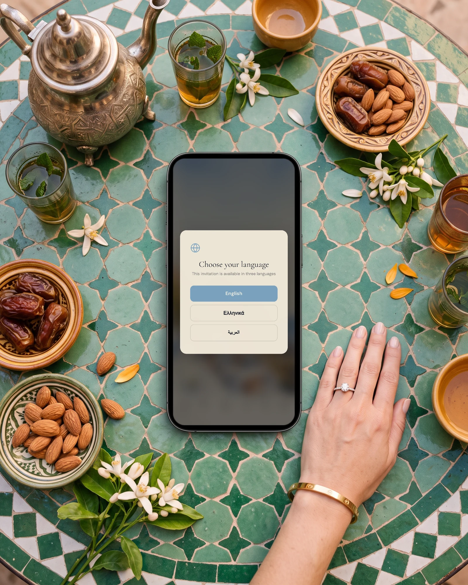 Nikah and walima invitation language choice on a phone, English, Greek or Arabic, lying on a zellige-tiled riad table with mint tea, a silver teapot, dates, almonds, orange blossom and a bride's hand