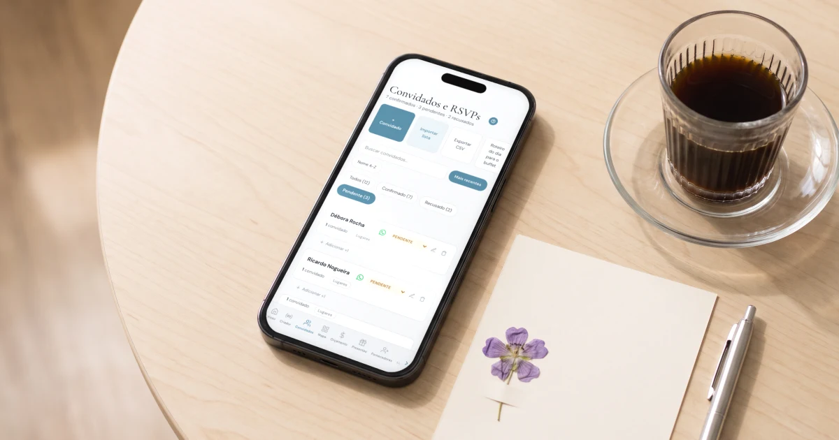 Site de casamento com confirmação de presença: celular deitado sobre uma mesa de madeira clara mostrando a lista de convidados filtrada nos pendentes, com o botão de WhatsApp em cada nome, ao lado de um save the date impresso e um cafezinho