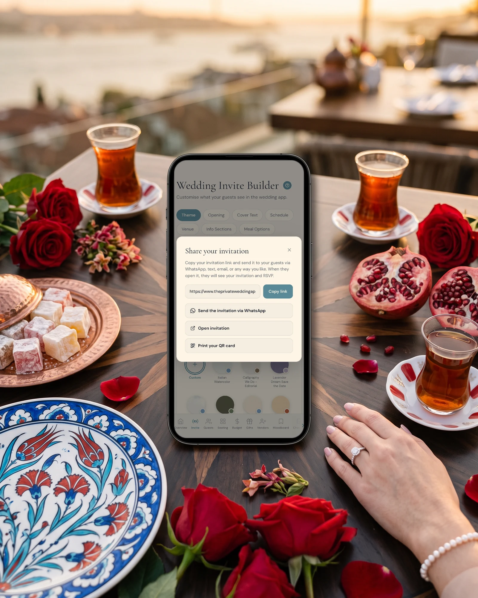 Turkish and English wedding invitation share panel on a phone, with Send the invitation via WhatsApp and Copy link, lying on a Bosphorus terrace table with Iznik plates, Turkish delight, pomegranates, tea glasses and red roses