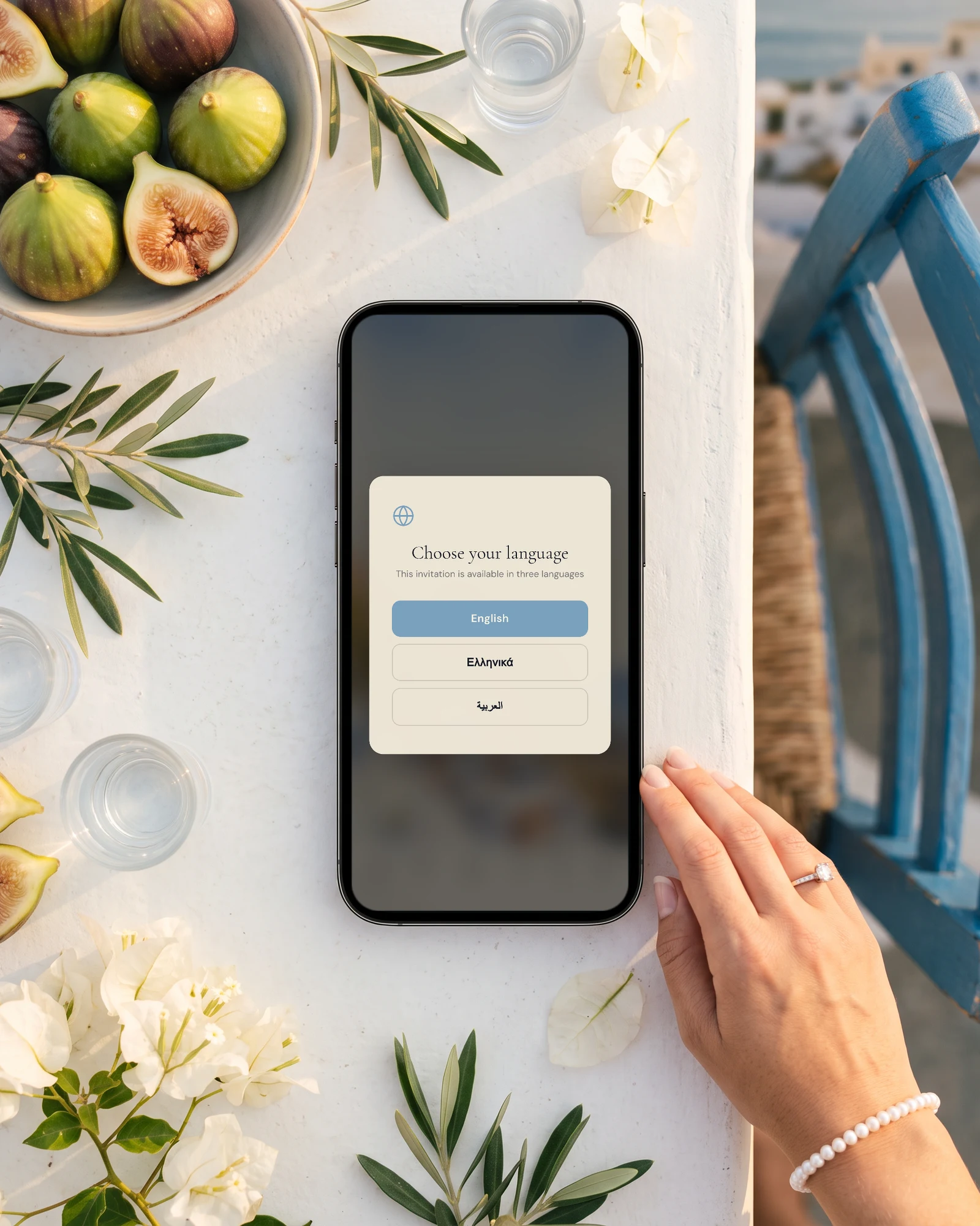Wedding abroad invitation with a language choice of English, Greek or Arabic on a phone lying on a white Greek island table, with figs, olive branches, glasses of ouzo, white bougainvillea and a bride's hand