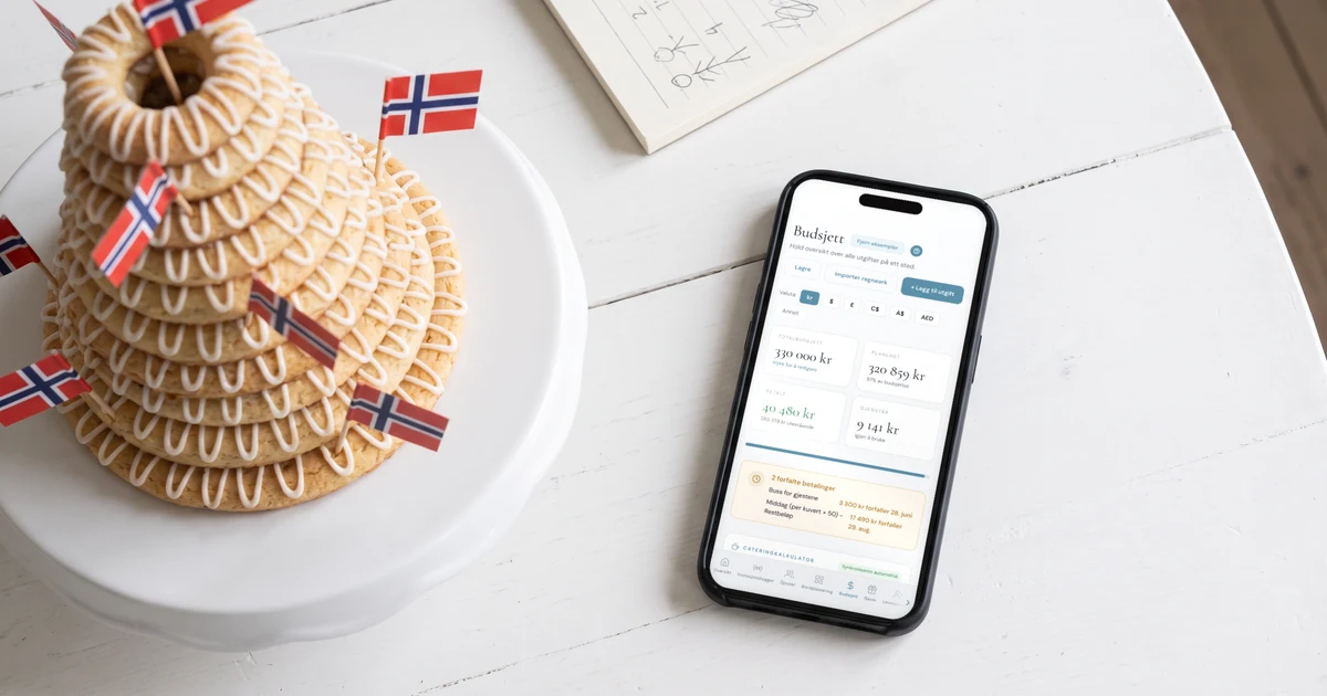 Kransekake med norske flagg på et kakefat ved siden av en mobiltelefon flatt på bordet som viser bryllupsbudsjettet med total, betalt og neste forfall, og en notatblokk