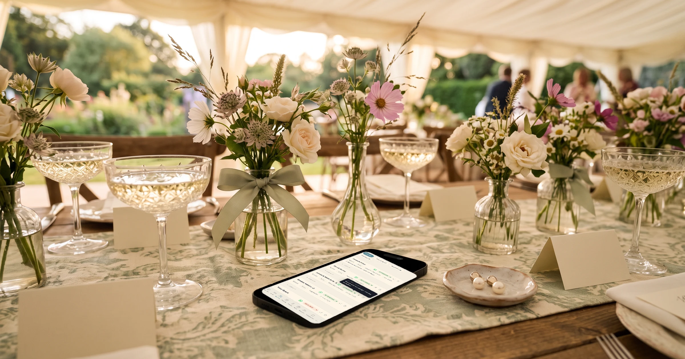Wedding invitation for a couple, two seats: wedding guest list with couples, plus-ones and meal choices on a phone, lying on a garden marquee table with wildflower bouquets, crystal coupes, a sage bow, blank place cards and pearl earrings