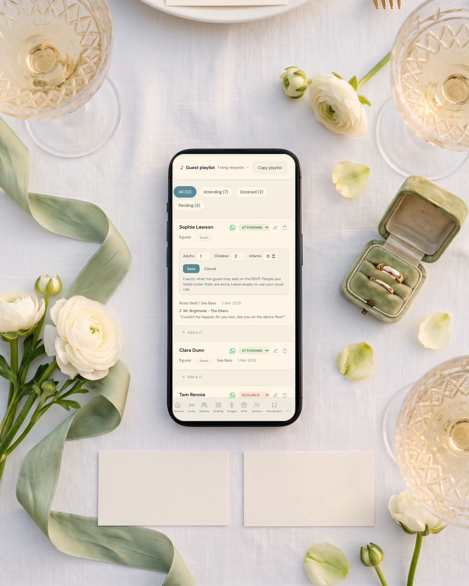 Wedding guest list on a phone setting seats per guest for adults, children and infants, lying on white linen beside two blank place cards, a sage silk ribbon, white ranunculus and a ring box with two wedding bands