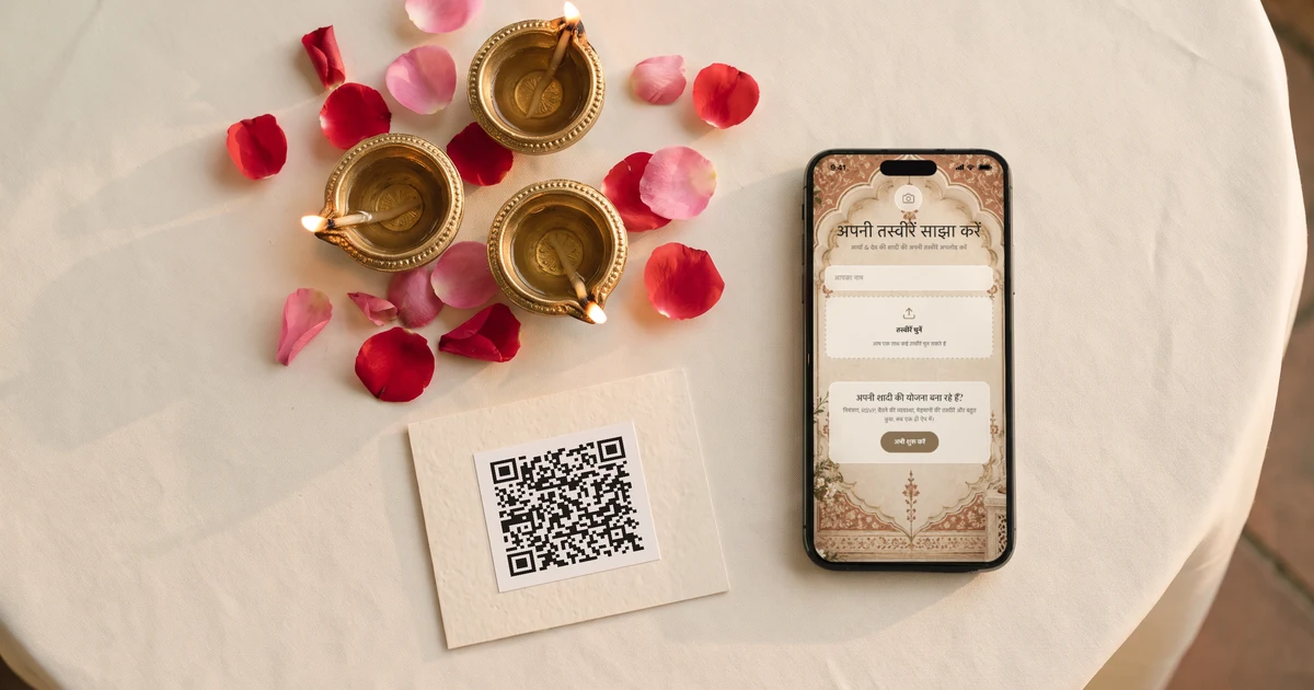 रिसेप्शन की गोल टेबल पर पीतल के दीये और गुलाब की पंखुड़ियों के बीच QR कोड वाला टेबल कार्ड, पास में मेज़ पर रखा फ़ोन जिसमें शादी की फ़ोटो अपलोड करने का पन्ना खुला है
