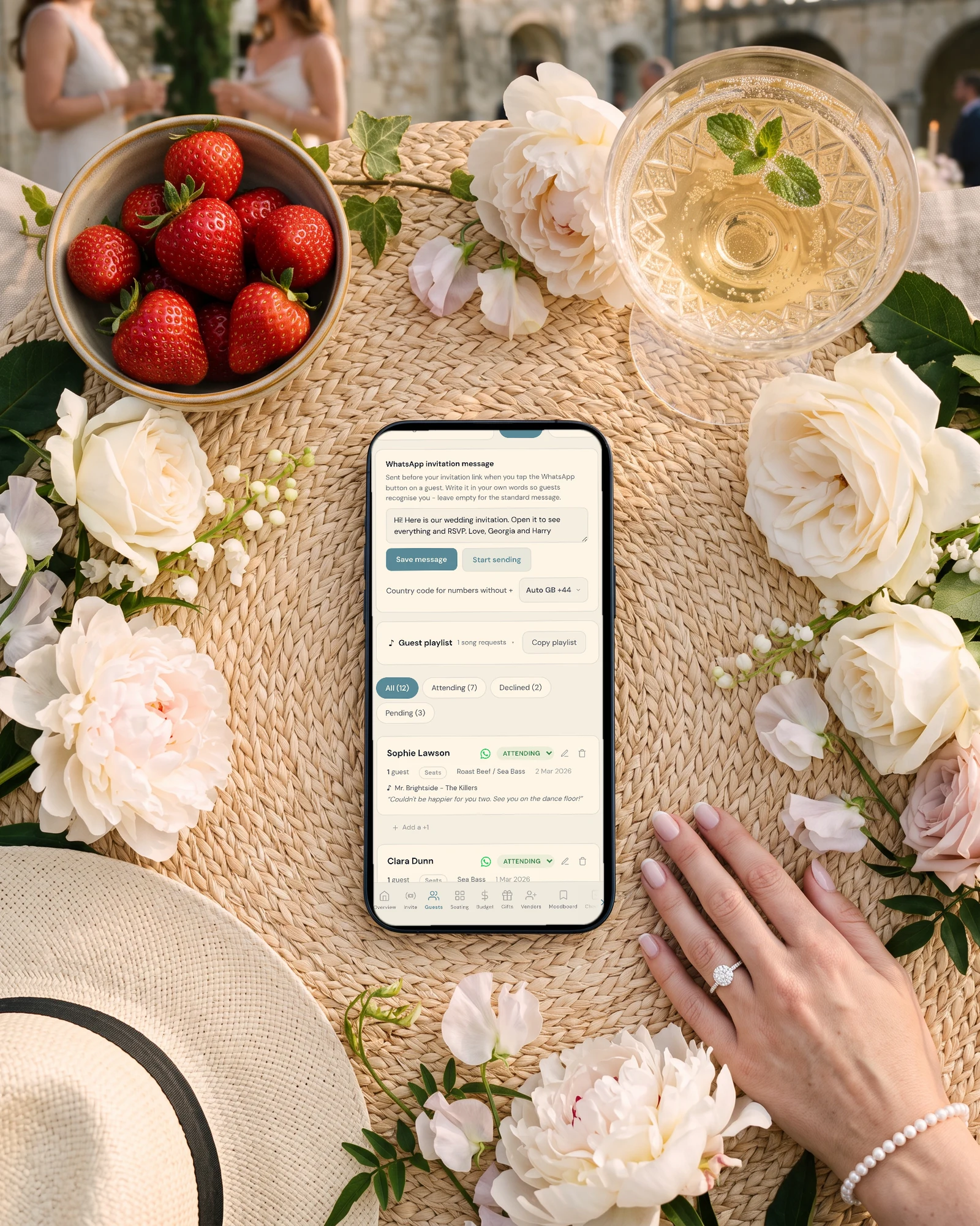 WhatsApp wedding invitation message card and guest list with WhatsApp buttons on a phone, for guests with only phone numbers, lying on a garden picnic mat with strawberries, peonies, a glass of lemonade and a bride's hand