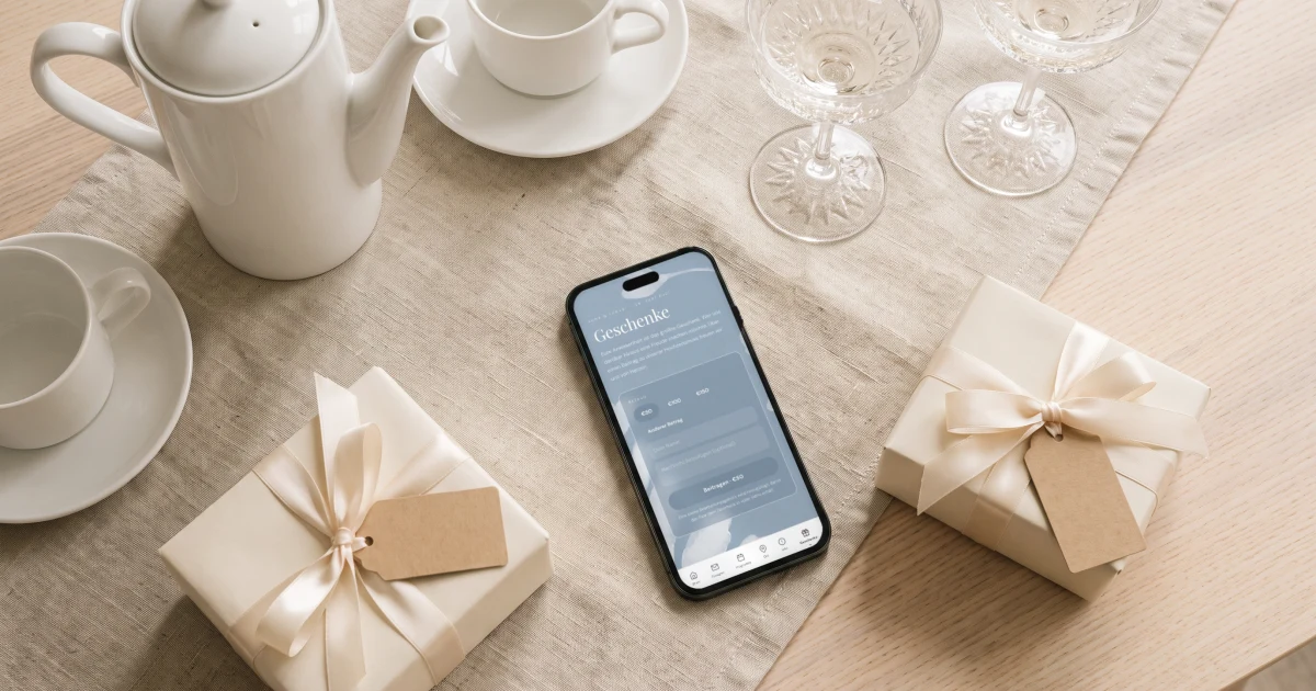 Wunschliste zur Hochzeit: ein Handy zeigt die Geschenkseite der Einladung neben Porzellan, Sektgläsern und einem Geschenk