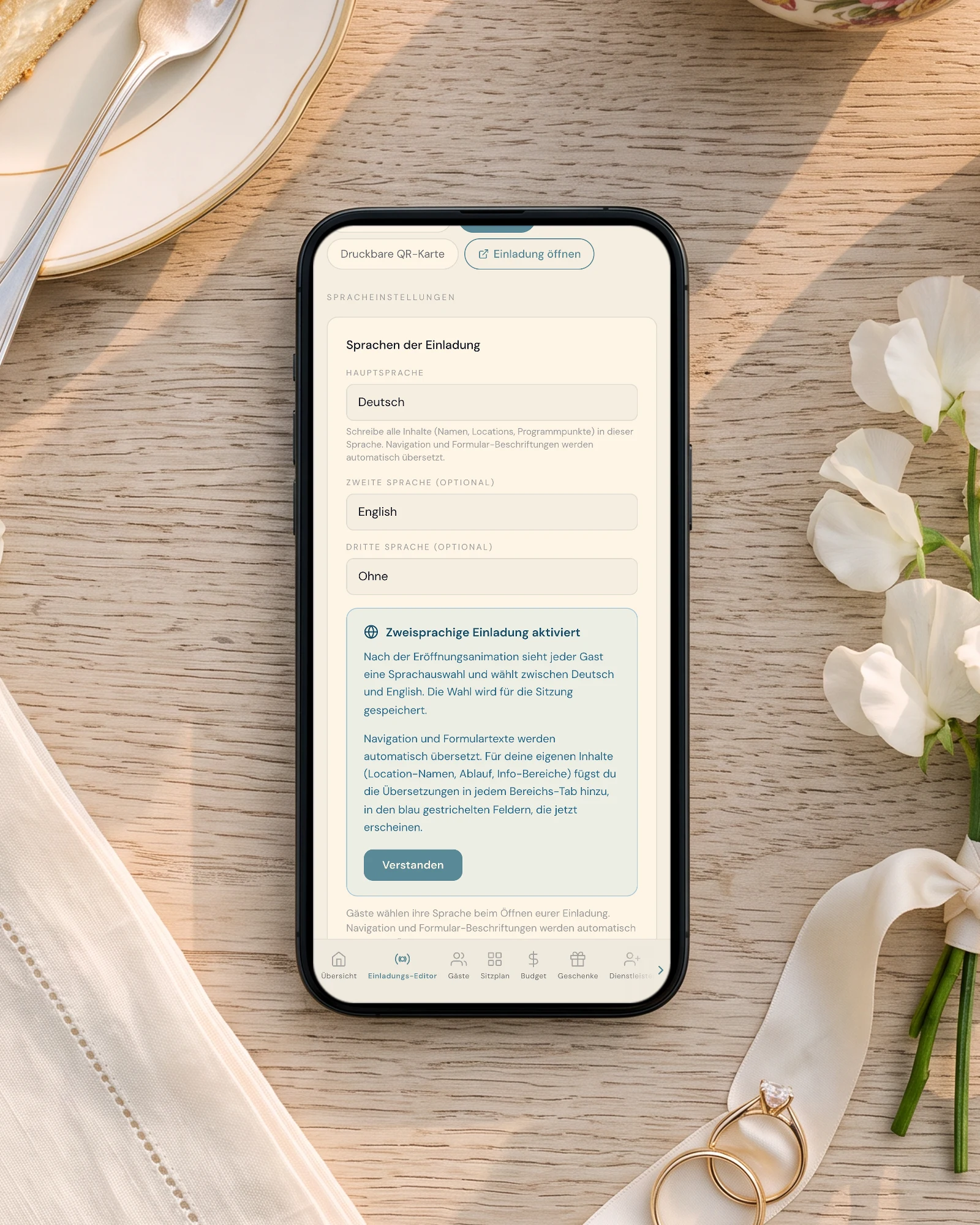 Zweisprachige Hochzeitseinladung Deutsch Englisch: Ein Handy auf einem Eichentisch neben Bienenstich, einer englischen Teekanne und Eheringen zeigt die Spracheinstellungen mit Hauptsprache Deutsch und zweiter Sprache English