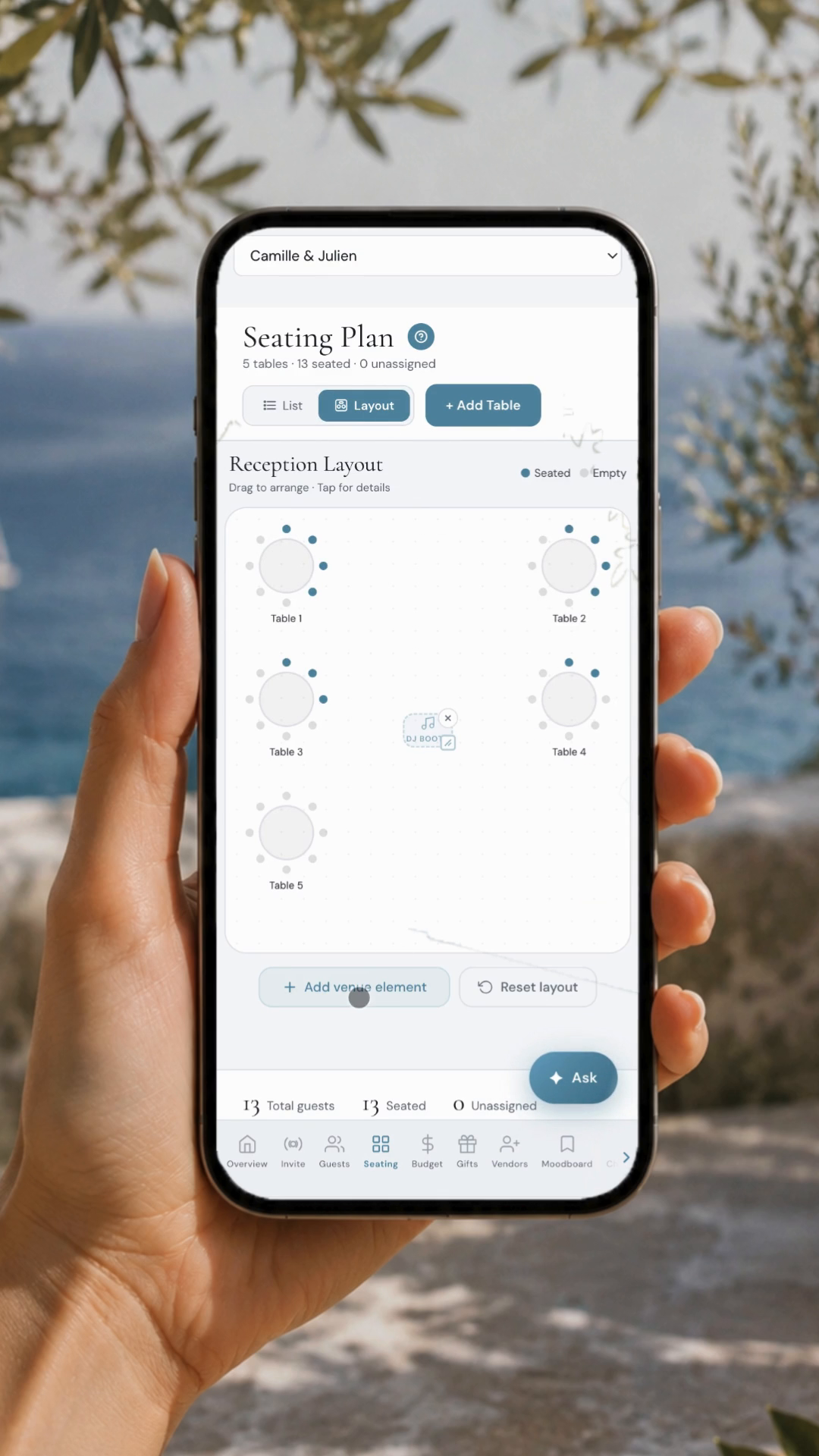 Wedding planner app behind the La Dolce Vita lemon invitation: drag-and-drop seating plan with reception layout and round tables, shown on iPhone against a Mediterranean coastline